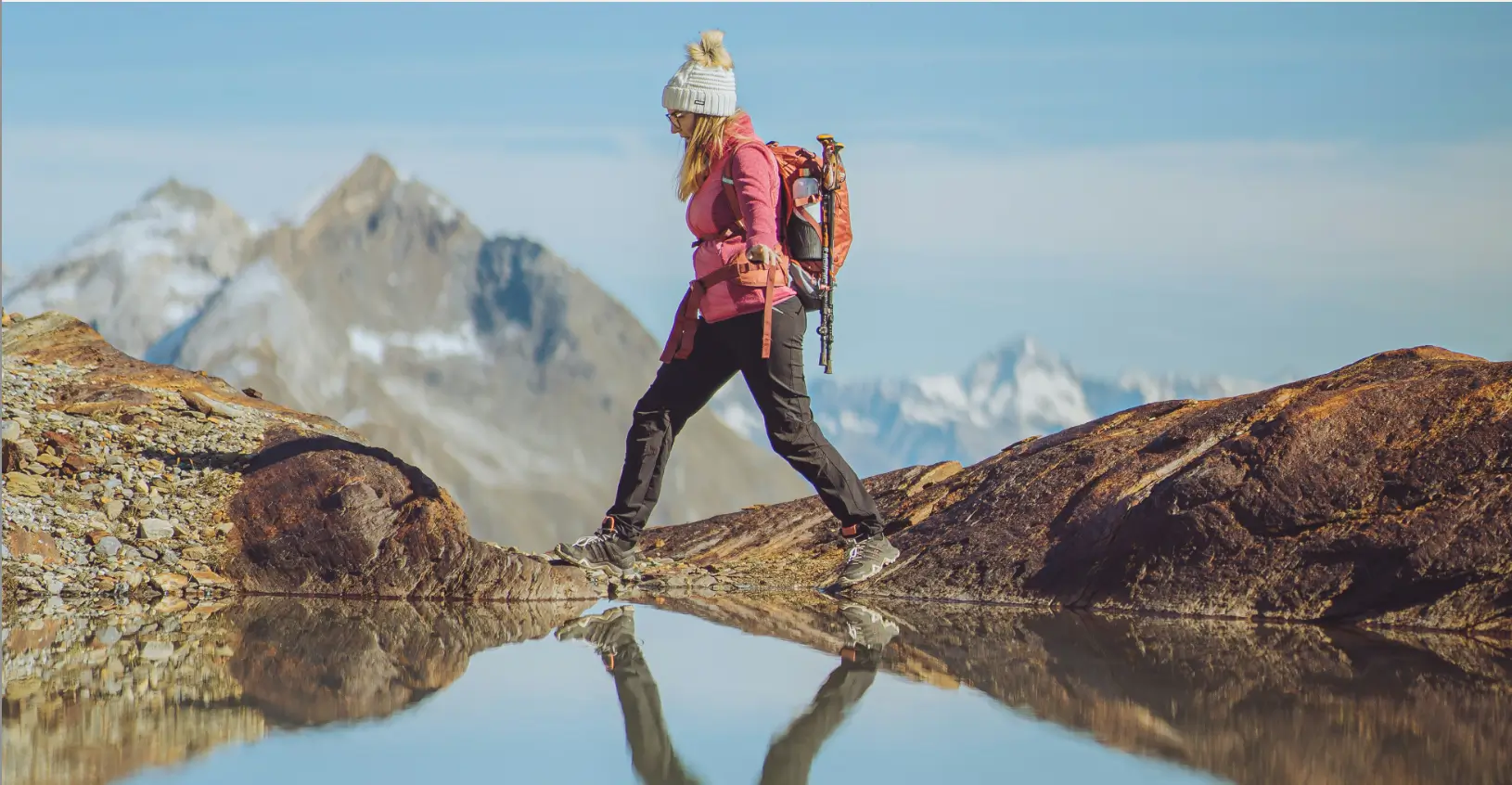 Hiker in a pink jacket crossing rocks above a still mountain lake